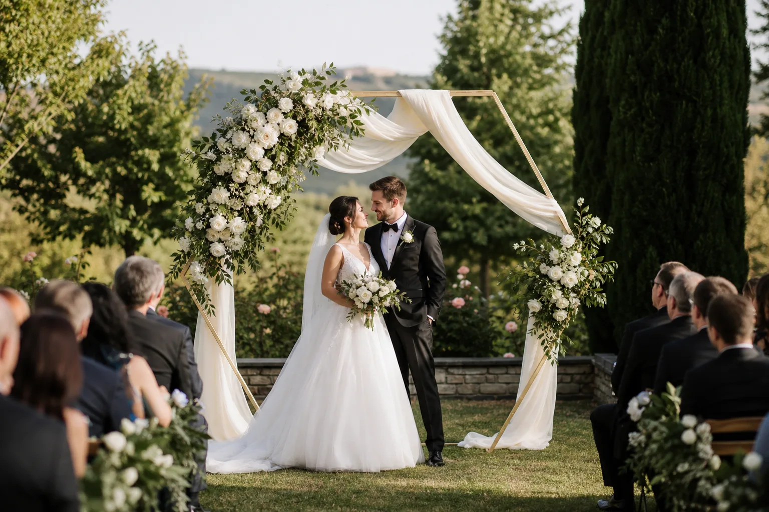 Tall champagne-gold hexagonal arch with ivory drape and white flowers at a full garden ceremony